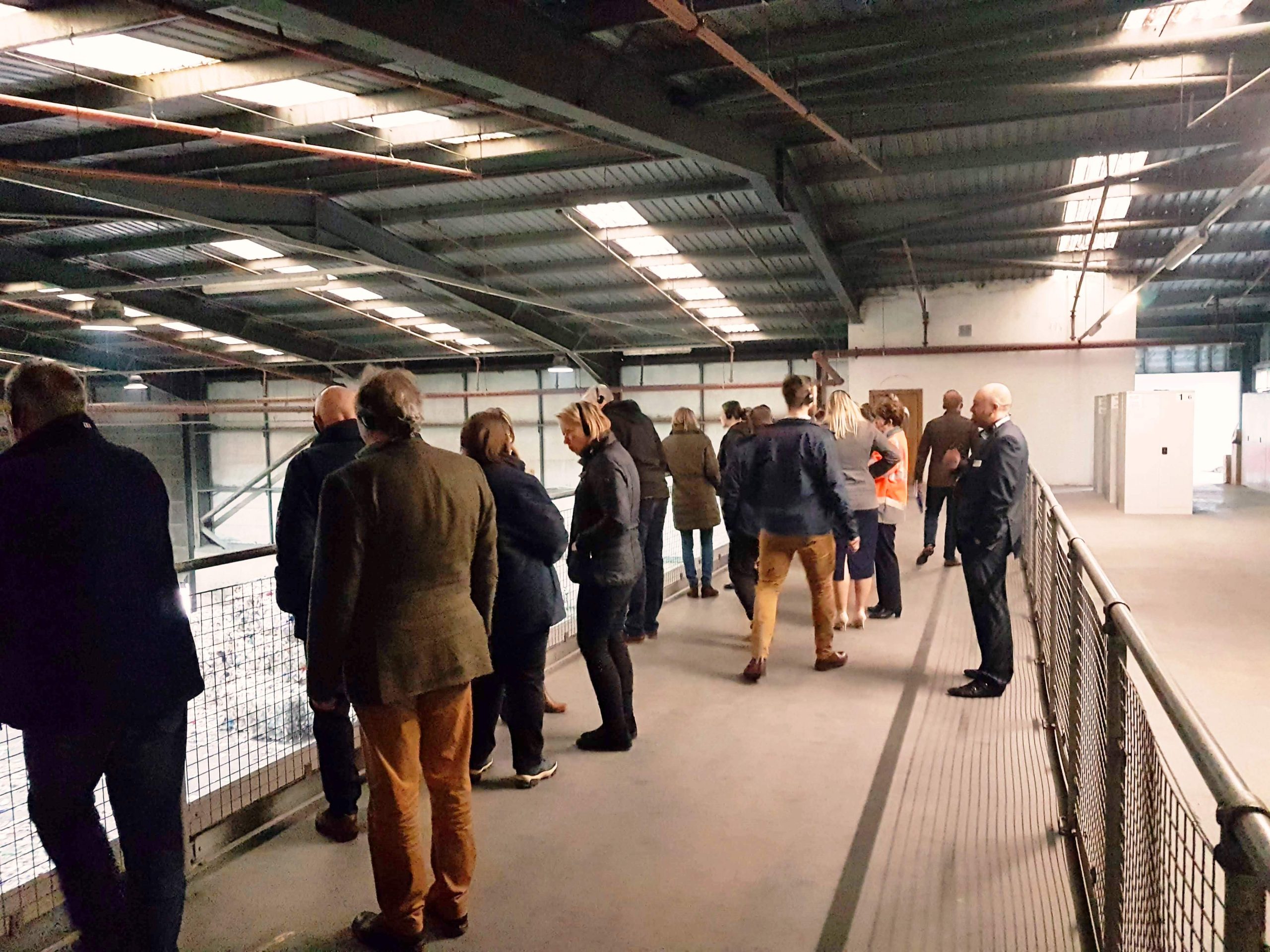 Image shows a floor area with low roof and handrails with members of the public looking over a factory floor.